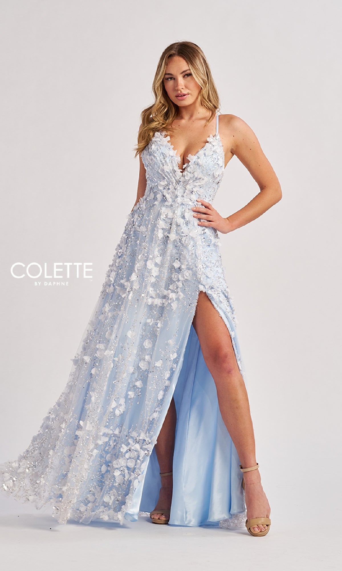 A woman stands against a plain background wearing the Colette CL8525, a light blue long prom dress with an A-line silhouette, embellished flowers, a thigh-high slit, and strappy heels.